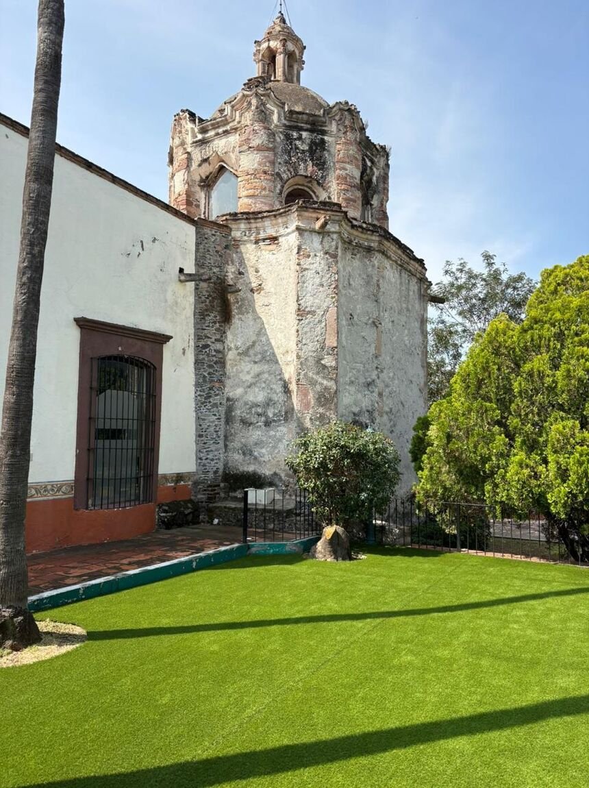 Jardín con pasto sintético Game Turf frente a una capilla de cantera, con remate de piedra y jardineras.