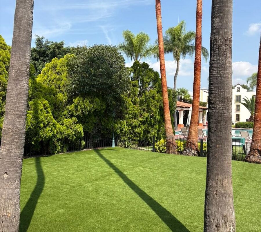 Jardín de pasto sintético instalado por Game Turf entre palmeras y arbolado, con la zona de alberca de un conjunto residencial al fondo.