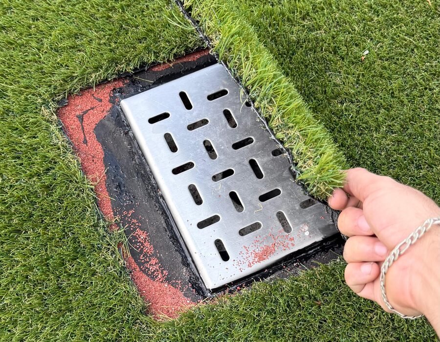 Mano levantando una tapa de pasto sintético que oculta la coladera de un registro, instalada por Game Turf.